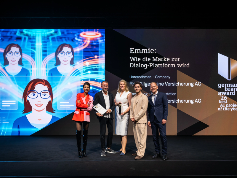 Sven Schmitt, Anke Josten und Eldin Thomas von der R+V Versicherung nehmen die Best-of-Best-Auszeichnung des German Brand Award 2026 für den KI-Chatbot Emmie entgegen, umrahmt von den Juroren Sophia Rödiger (links) und Christian Rummel (rechts).