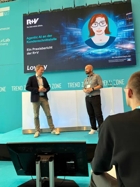 Ulf Loetschert von LoyJoy und Peter Paul Nasim von der R+V Versicherung halten einen gemeinsamen Vortrag auf der Bühne der insureNXT in der Trend Zone. Im Hintergrund ist eine große Leinwand mit der Präsentation 'Agentic AI an der Kundenschnittstelle' zu sehen.