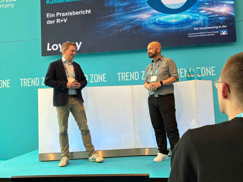 Ulf Loetschert von LoyJoy und Peter Paul Nasim von der R+V Versicherung halten einen gemeinsamen Vortrag auf der Bühne der insureNXT in der Trend Zone. Im Hintergrund ist eine große Leinwand mit der Präsentation 'Agentic AI an der Kundenschnittstelle' zu sehen.